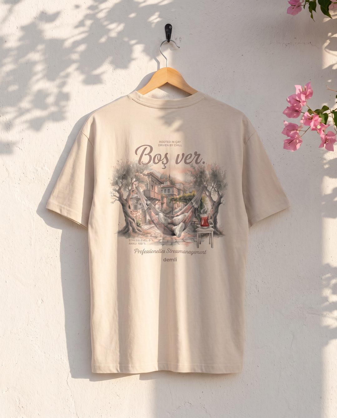 demli Boş ver. Oversized T-Shirt in Sand auf einem Holzbügel an einer weißen Kalkwand mit Bougainvillea