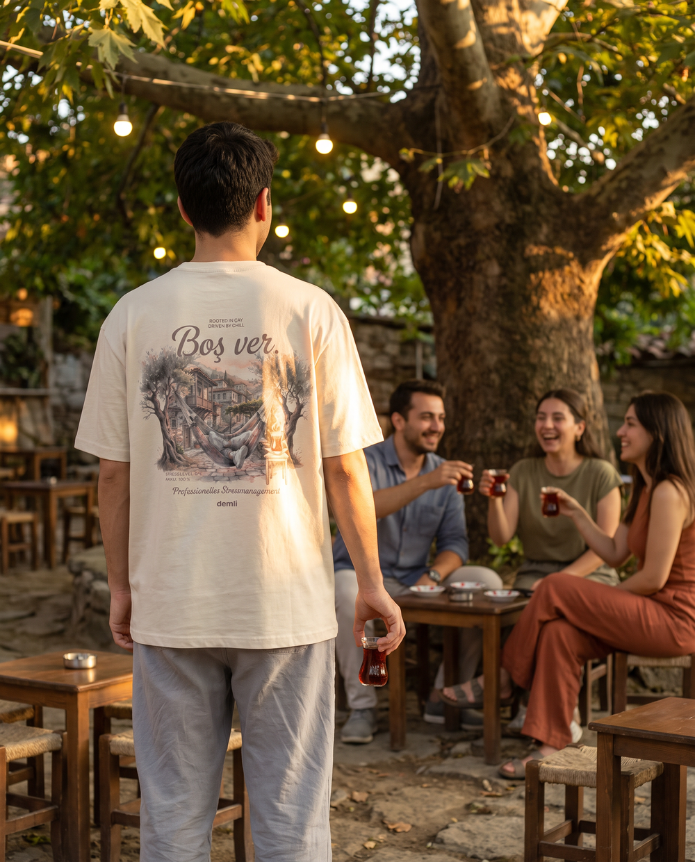 Person im demli Boş ver. Shirt in Sand von hinten im Teegarten, Freunde im Hintergrund