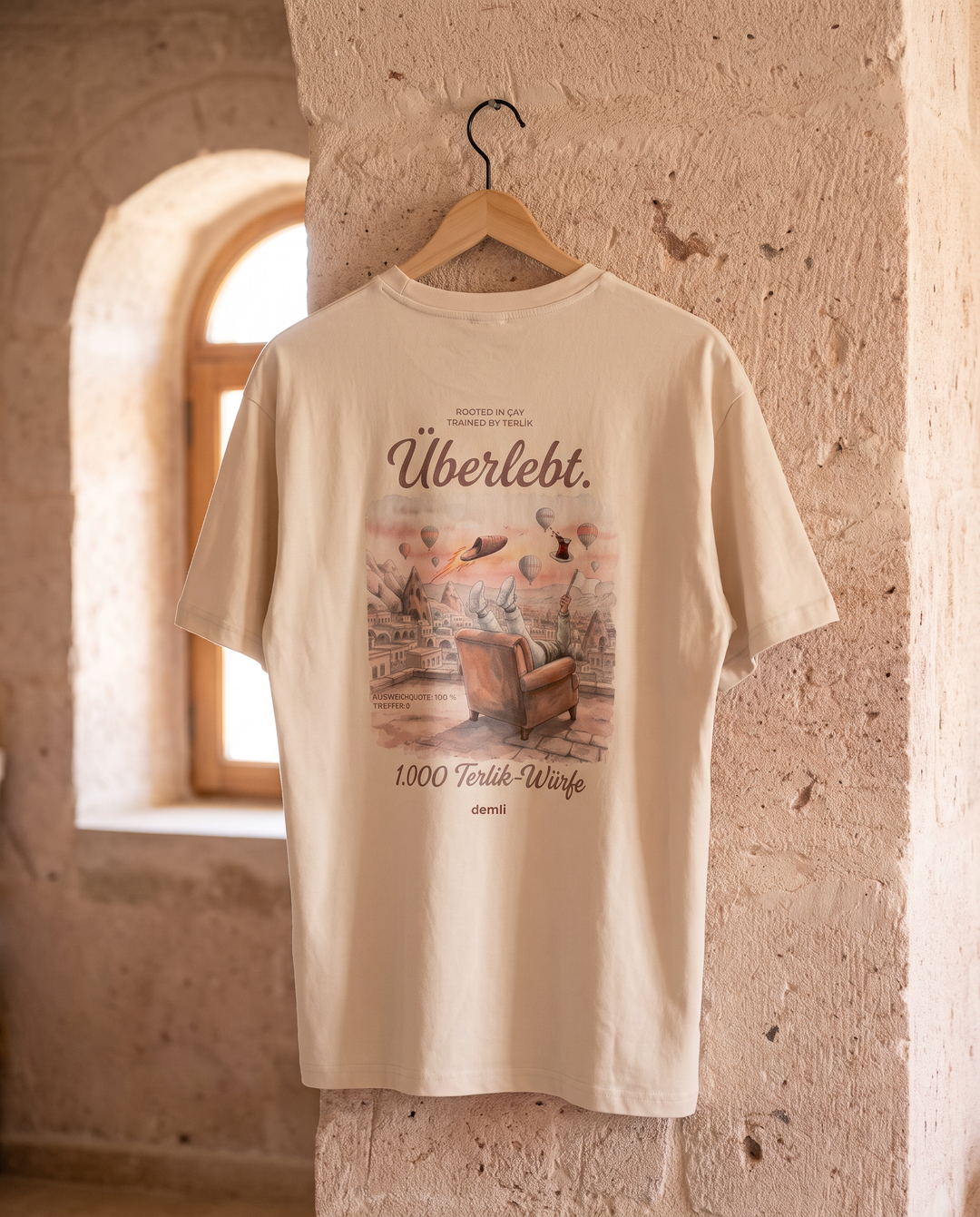demli Überlebt. Oversized T-Shirt in Sand auf einem Holzbügel an einer Tuffsteinwand in Kappadokien